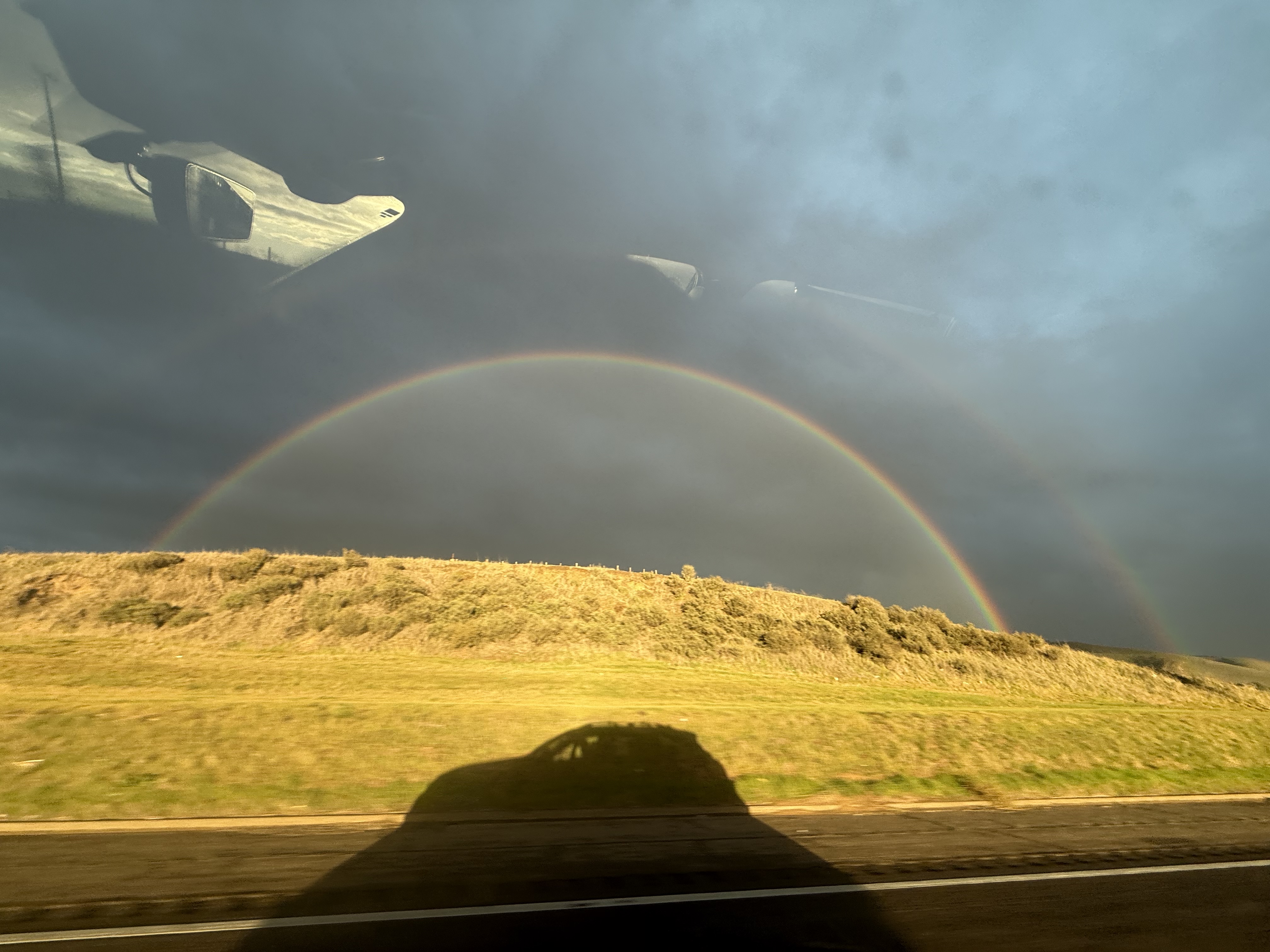 Full rainbow against a cloudy sky, with a car's shadow stretching into the grass on the side of the highway